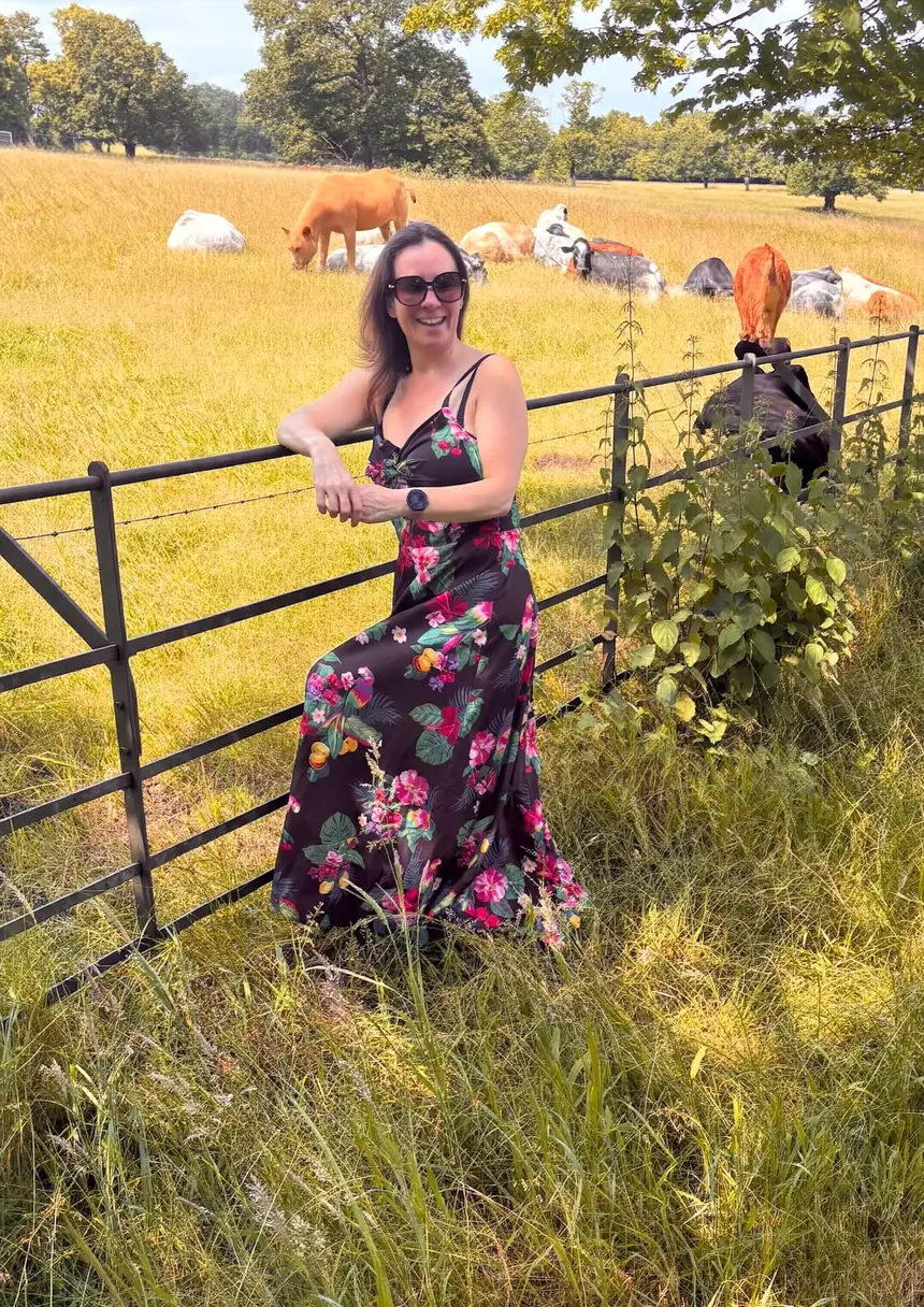 Lynette Tidy smiling, leaning on a field gate with cattle grazing behind her